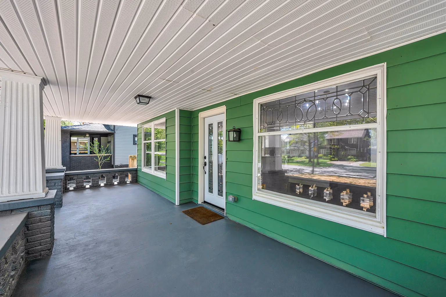 Spacious green house front porch with white door, large windows, and gray floor under white ceiling.
