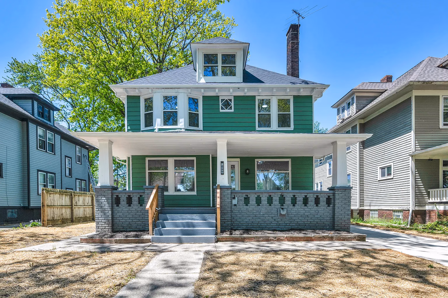 Two-story green house with white trim, large porch, brick pillars, and a chimney under a clear blue sky.