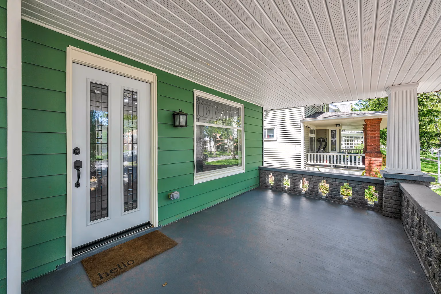 Empty covered front porch with green siding, white door with glass panels, and a brown doormat saying hello.