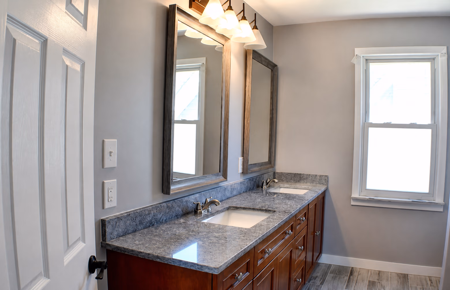 Bathroom with double sink vanity, gray granite countertop, wooden cabinets, and a window on the right wall.