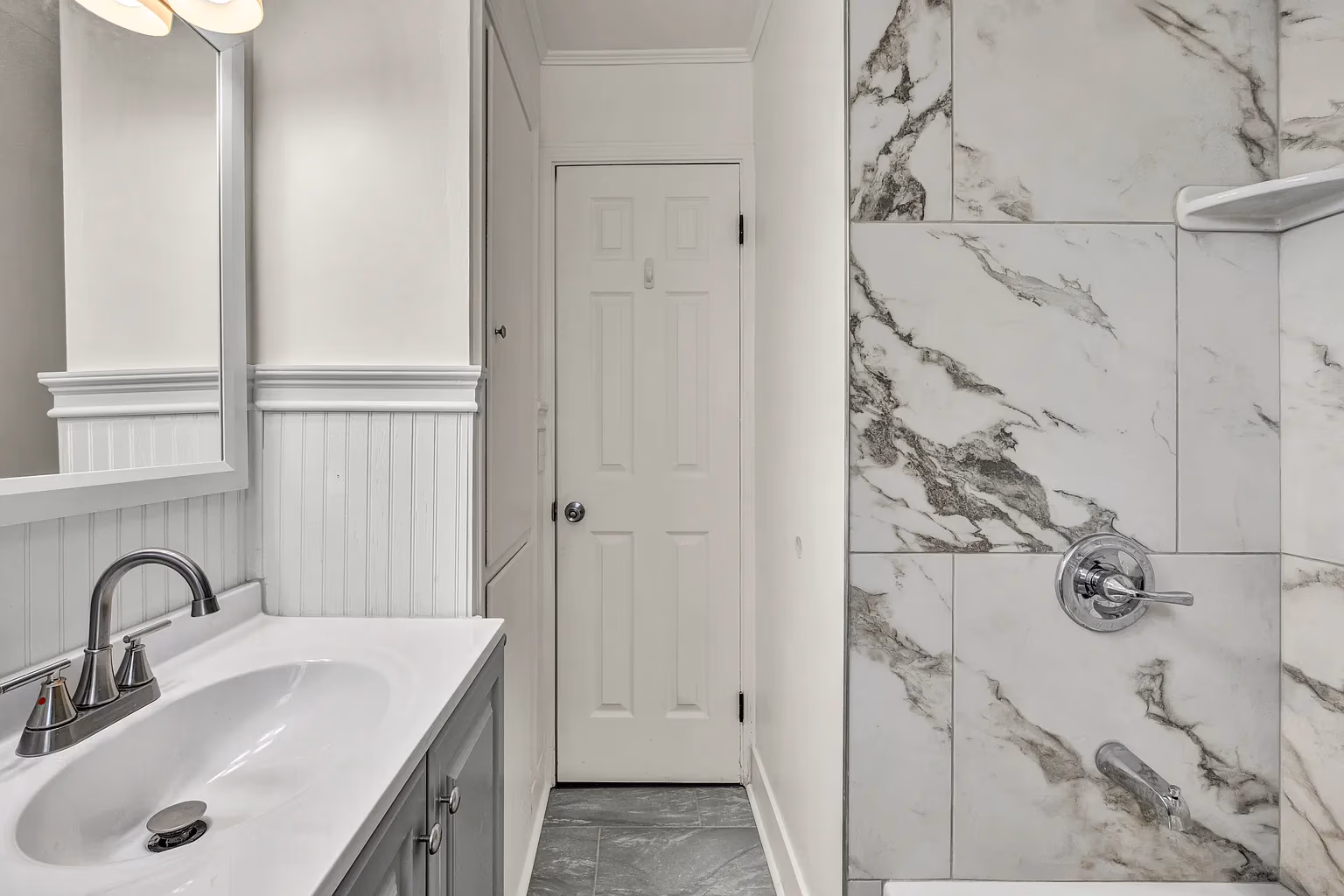 Modern bathroom with white sink, mirror, and a shower with gray marble tiles and chrome fixtures.