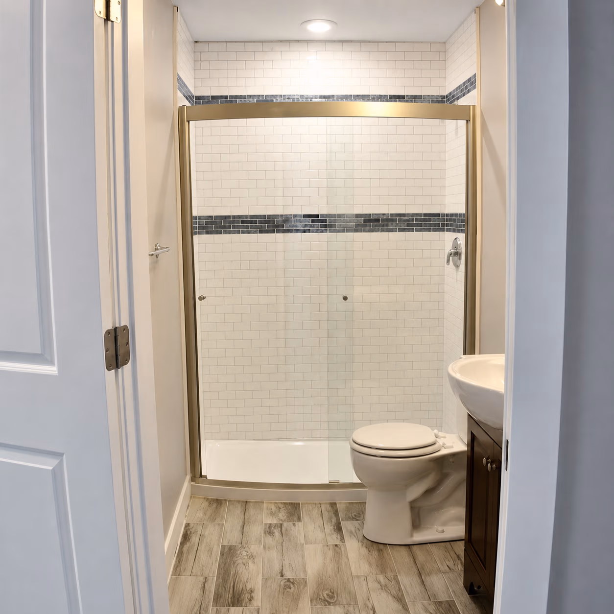 Small bathroom with wood-look tile floor, white toilet, sink, and glass shower with tiled walls.