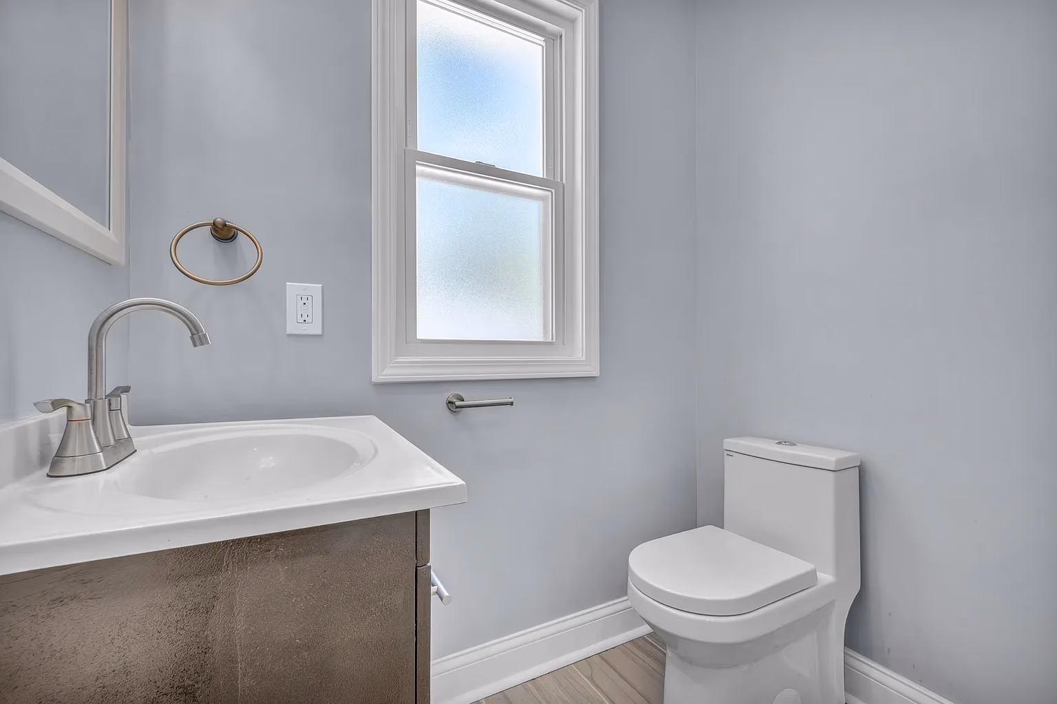 Minimalist bathroom with white toilet, sink, silver faucet, towel ring, and frosted window on gray walls.