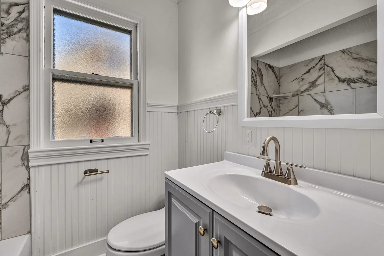 Compact bathroom with gray vanity, white sink, frosted window, and marble tile shower walls.