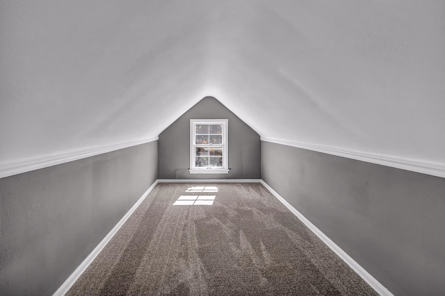 Empty attic room with slanted white ceiling, gray walls, carpeted floor, and a window at the far end.
