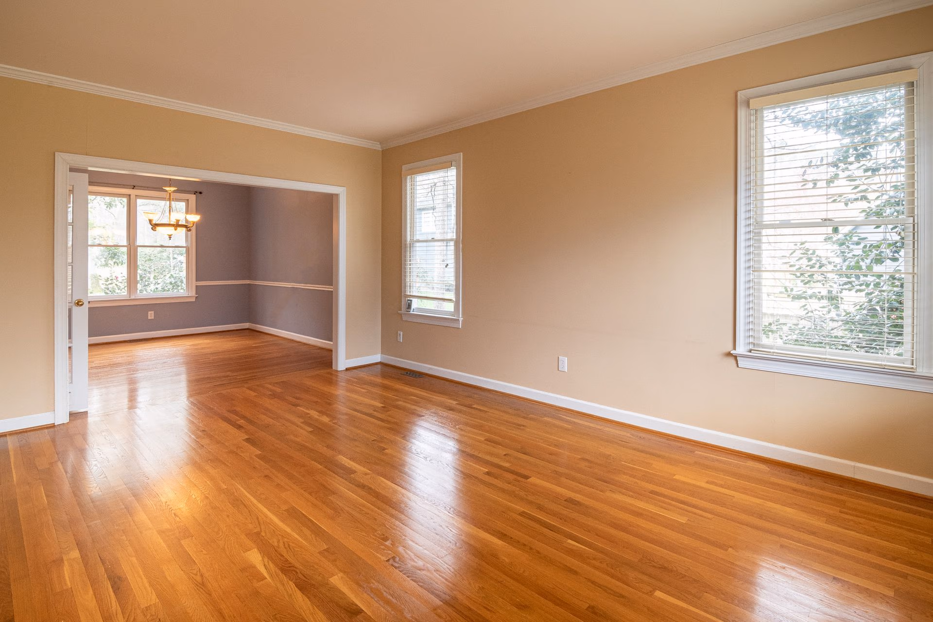 Empty room with polished hardwood floors, beige walls, two windows, and an adjacent dining area with chandelier.