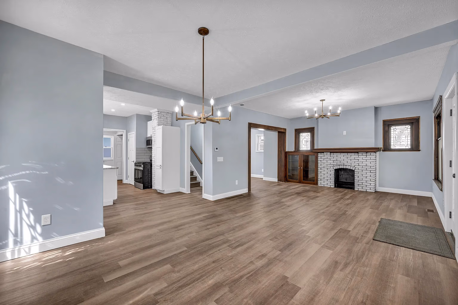 Spacious living room with light wood flooring, gray walls, a white brick fireplace, and two chandeliers.