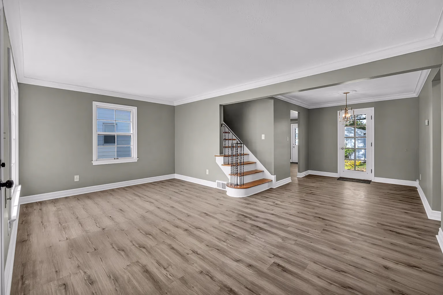 Empty living room with light gray wood flooring, gray walls, white trim, staircase, and glass door.