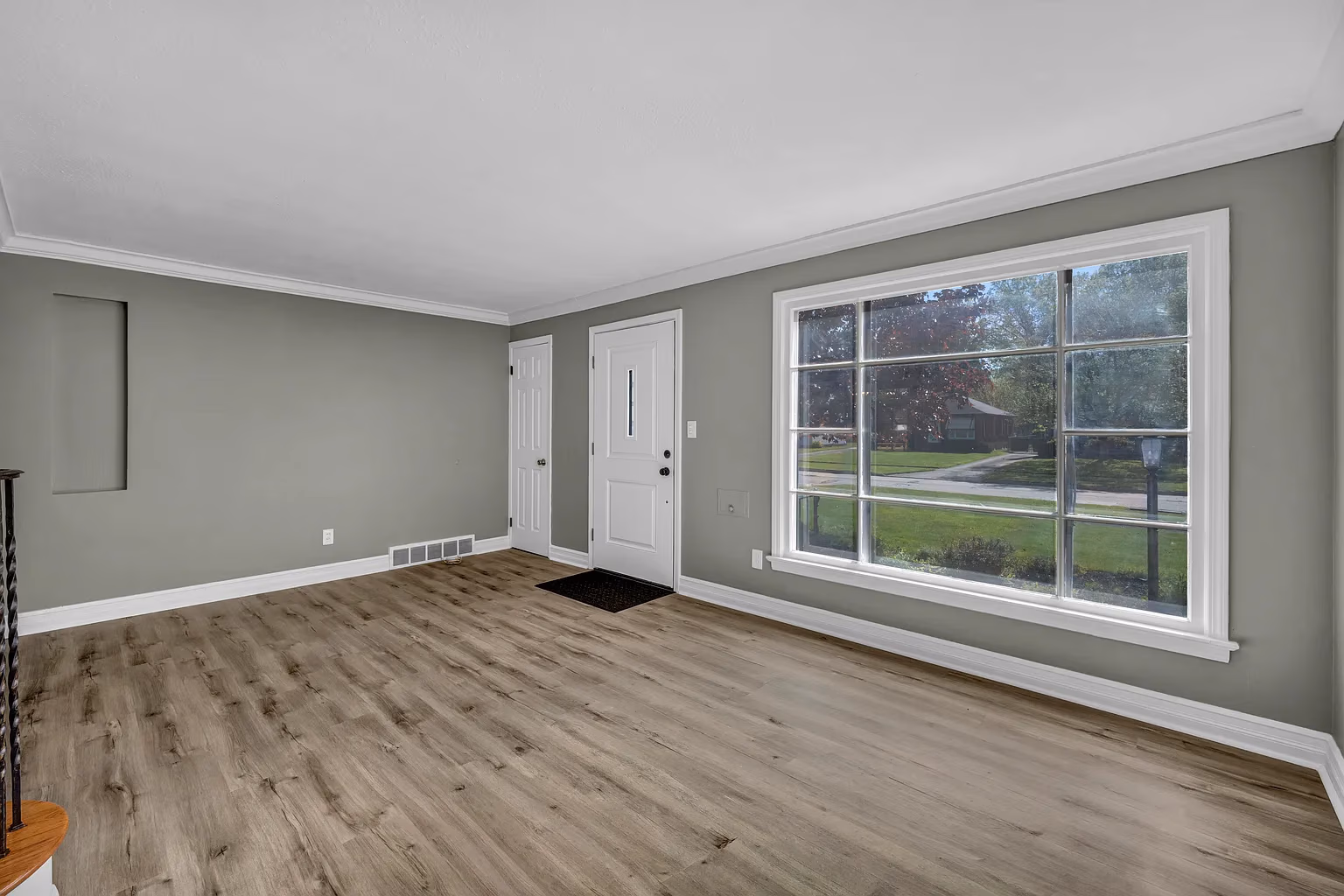 Empty room with gray walls, light wood flooring, white door, and large window showing front yard.