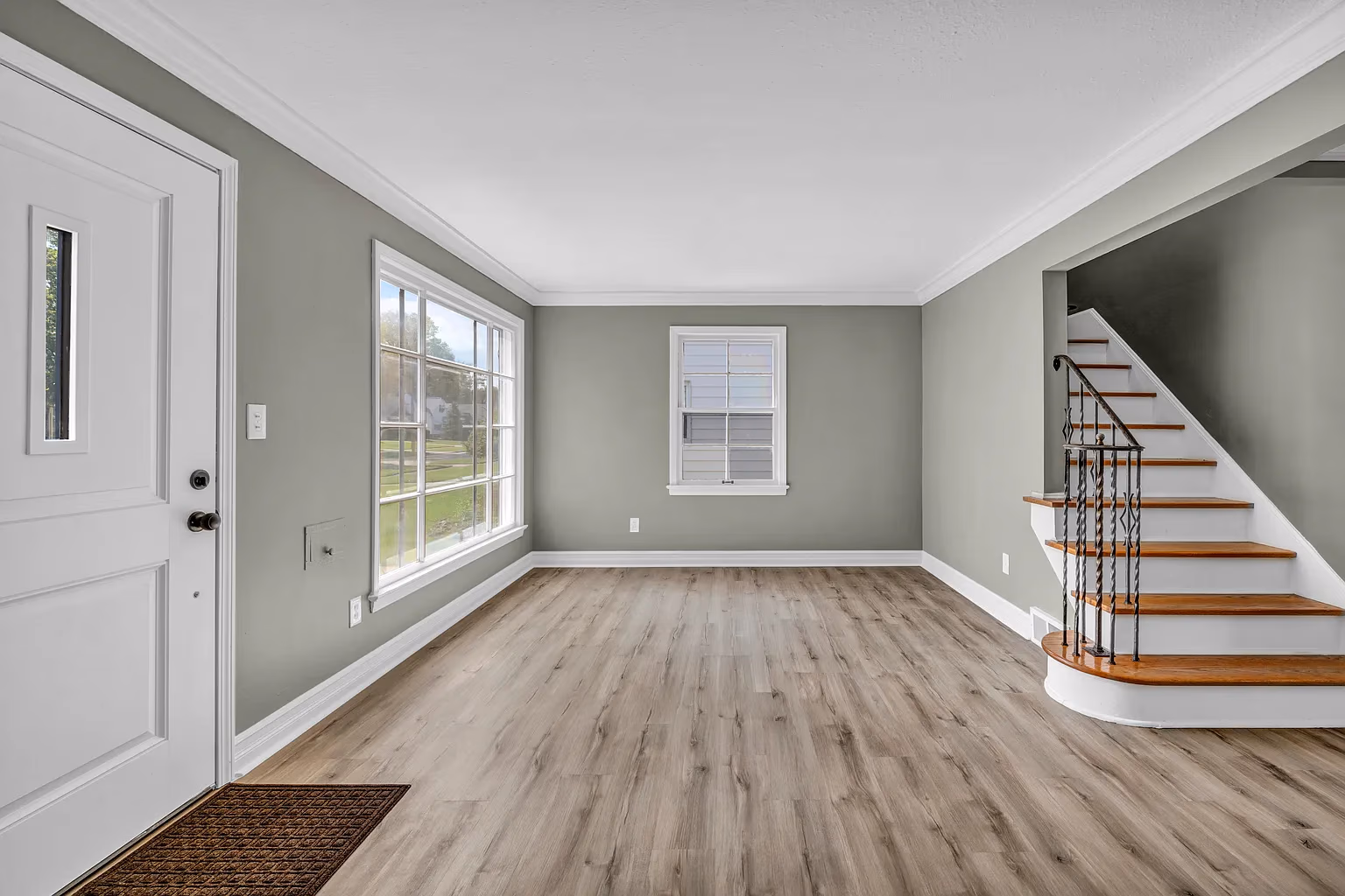 Empty room with light wood flooring, gray walls, white door, large window, and wooden stairs with iron railing.