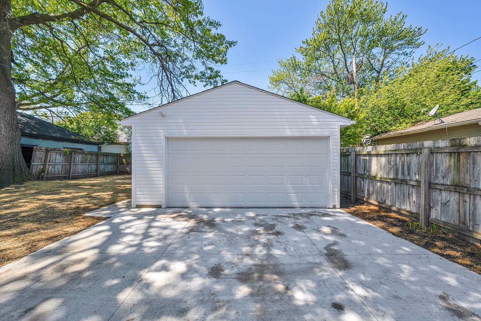 White detached double garage with closed door and concrete driveway surrounded by wooden fence and green trees.