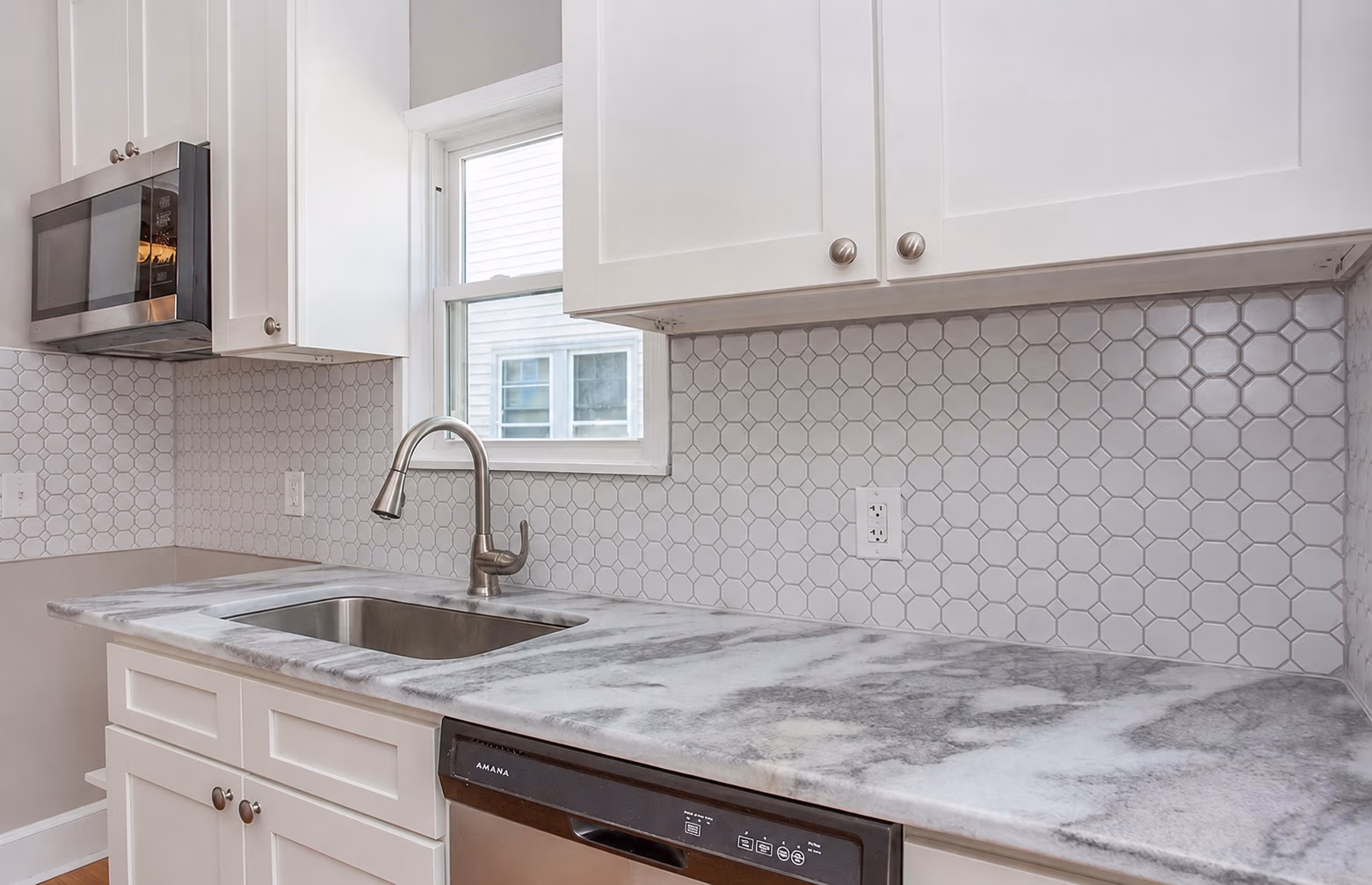 Modern kitchen countertop with marble surface, steel sink, faucet, white cabinets, and hexagonal tile backsplash.