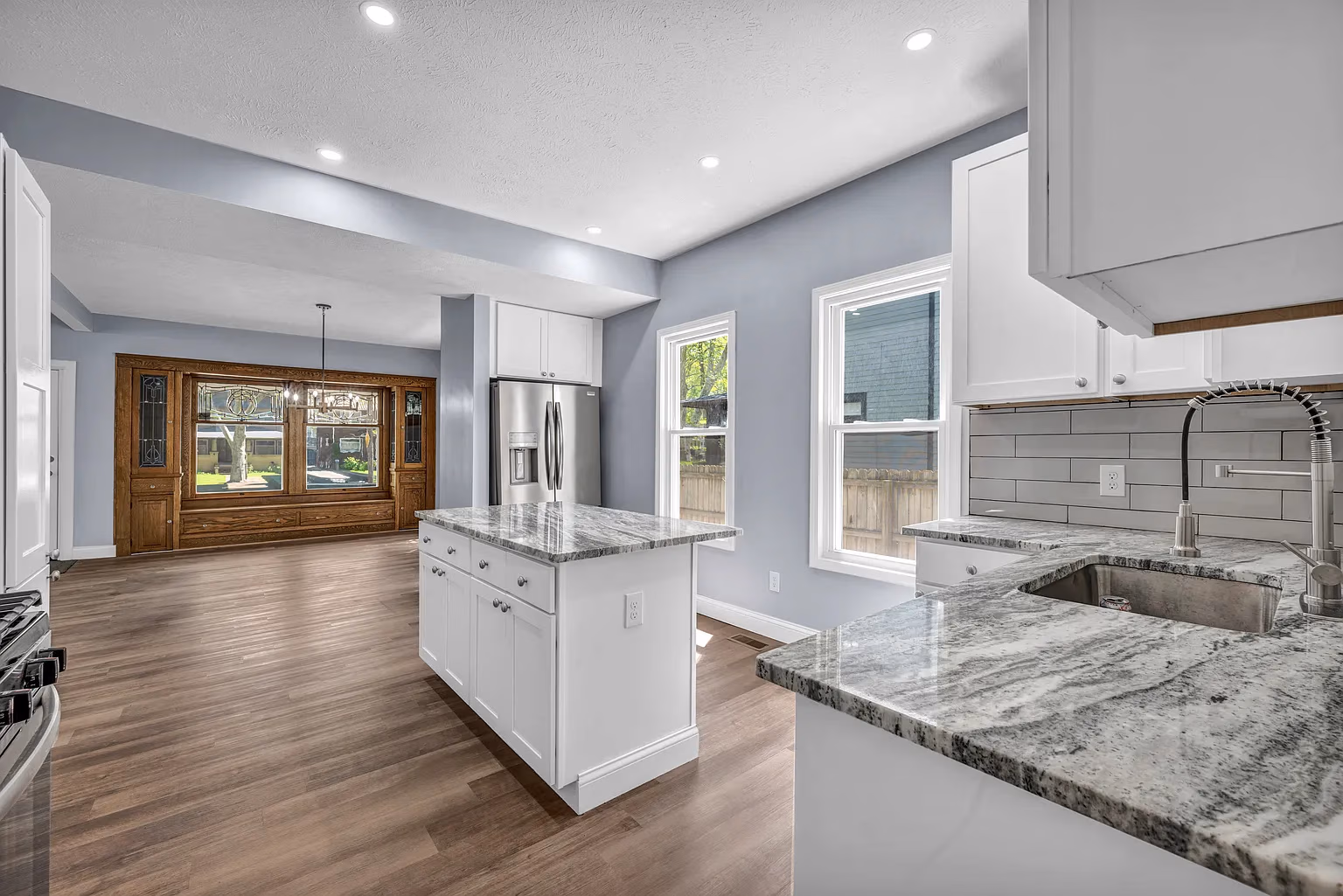 Modern kitchen with white cabinets, marble countertops, stainless fridge, and wood floors under recessed ceiling lights.