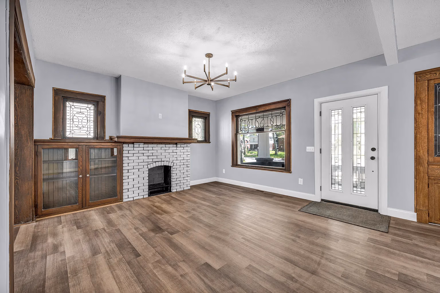 Empty room with gray walls, wood floor, white brick fireplace, stained glass windows, and white front door.