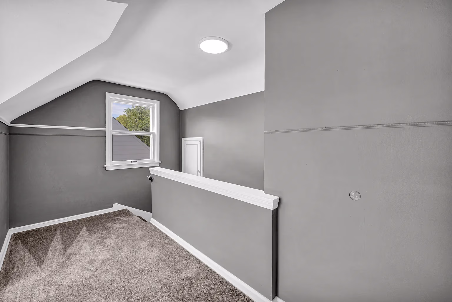 Empty gray room with carpeted floor, half-wall railing, window, and small white door under sloped ceiling.