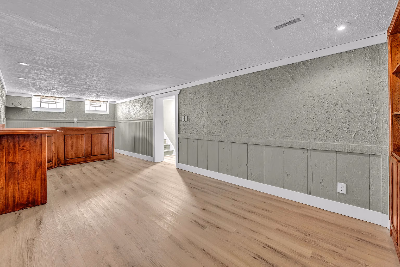 Empty basement room with light wood flooring, textured green walls, stairway, and built-in wood bar and shelves.