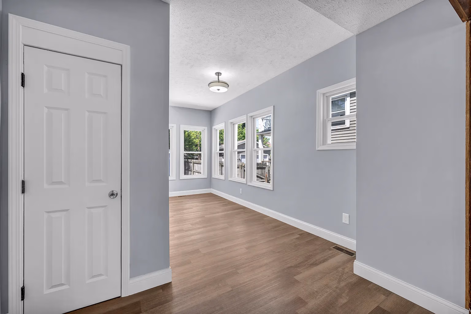 Empty room with light gray walls, wooden floor, white door, and several windows with outdoor view.