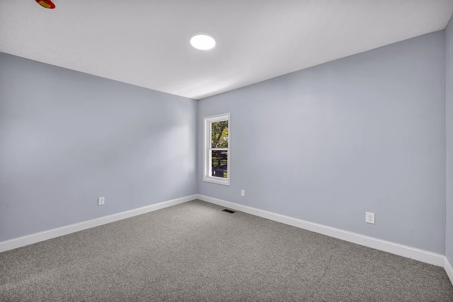 Empty room with light gray carpet, light blue walls, a small window, and a ceiling light.