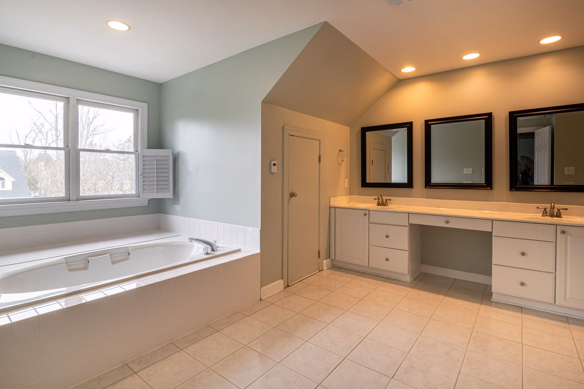 Spacious bathroom with large white bathtub, three mirrors, double sinks, and tiled floor.