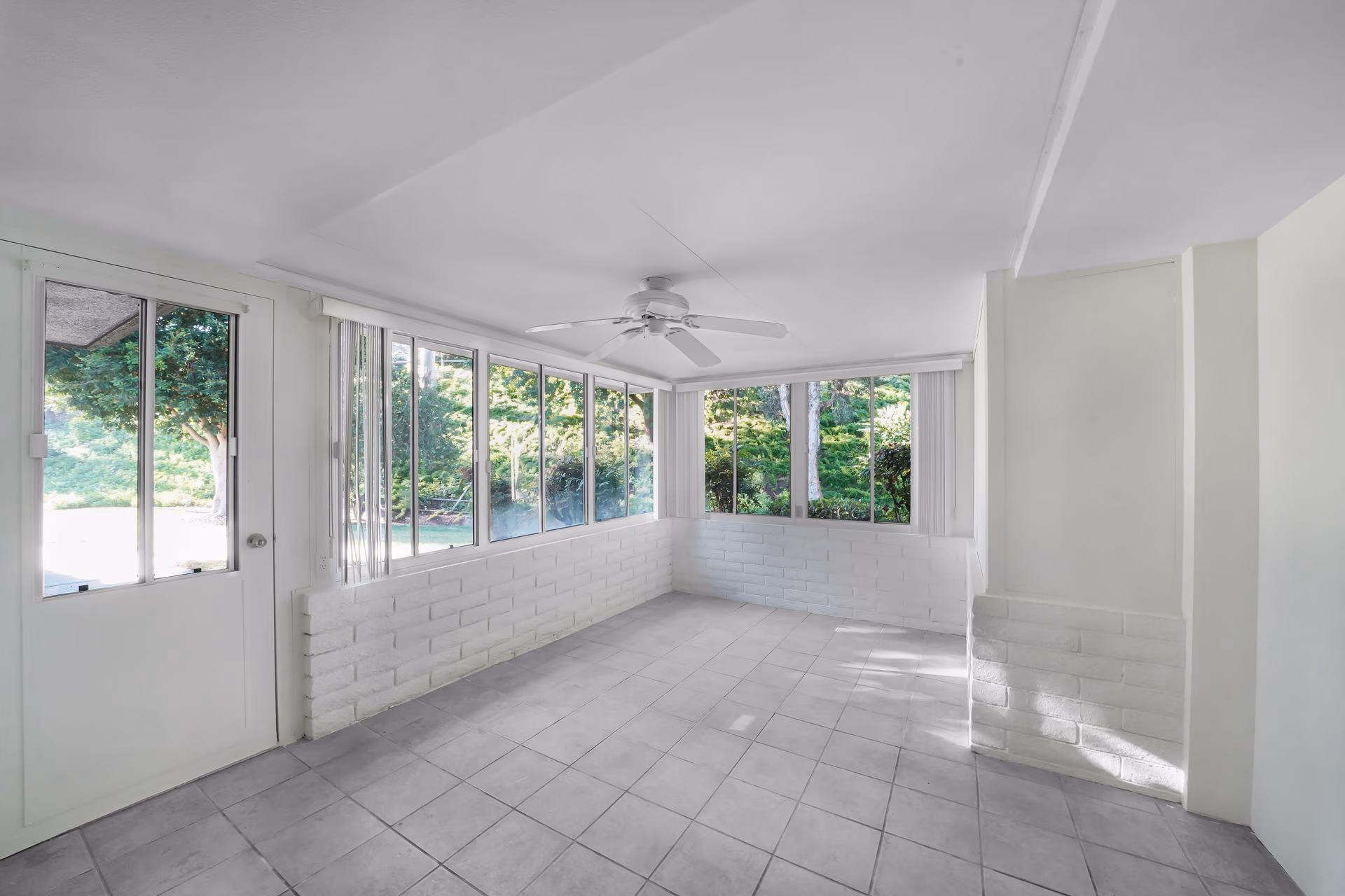 Empty sunroom with large windows, white walls, tiled floor, and ceiling fan overlooking a garden.