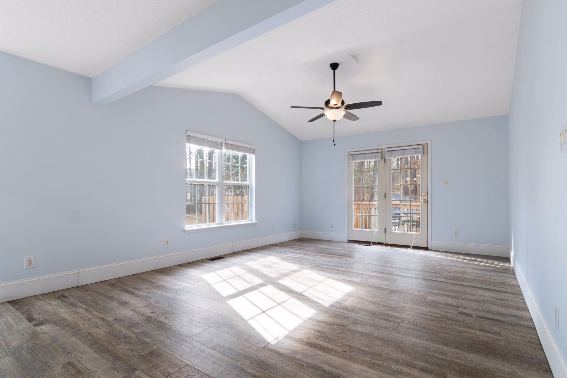 Empty room with light blue walls, wood flooring, ceiling fan, window, and glass door leading outside.